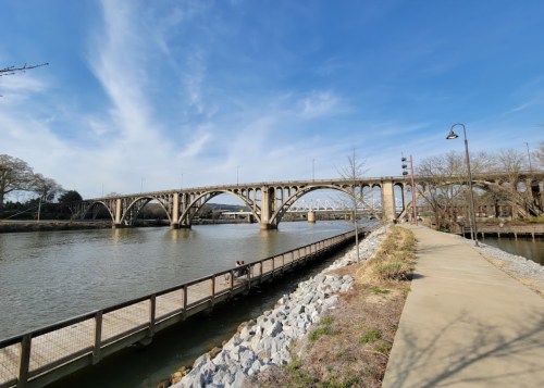 A concrete bridge spans a river, with a pathway and rocky shoreline visible under a clear blue sky.