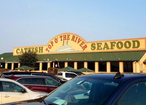 Sign for "Catfish Top O' The River Seafood" restaurant with a blue sky in the background and parked cars in the foreground.