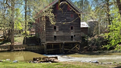 Historic wooden mill beside a calm river, surrounded by trees and greenery, with a small dam in the foreground.