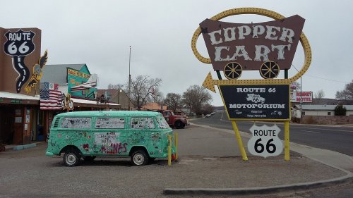 A vintage green van parked near a large sign for the Copper Cart on Route 66, with shops and flags in the background.