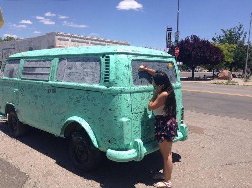 A person writes on a turquoise van covered in signatures and drawings, set against a sunny street scene.