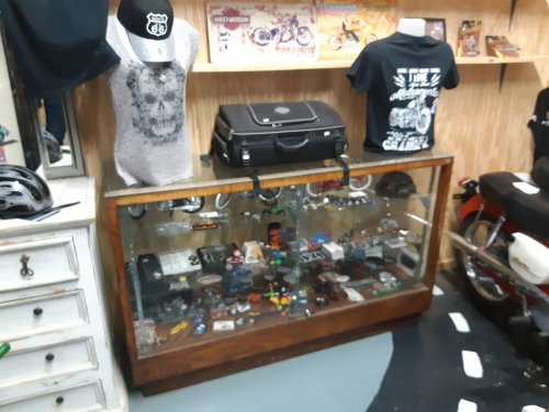 Display case filled with collectibles, clothing, and accessories in a shop, with a motorcycle in the background.