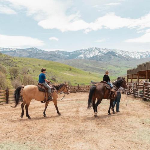 Two riders on horseback in a scenic outdoor setting with mountains in the background and a wooden fence nearby.
