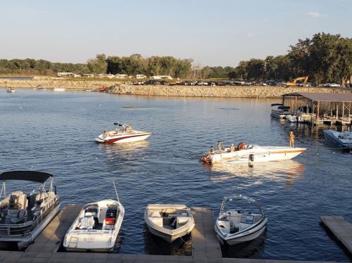 Boats on a calm lake with people enjoying the water, surrounded by trees and a rocky shoreline.