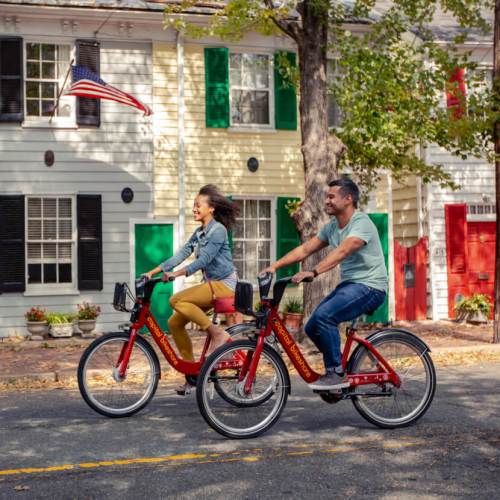 Two people ride bicycles on a tree-lined street with colorful houses and an American flag in the background.