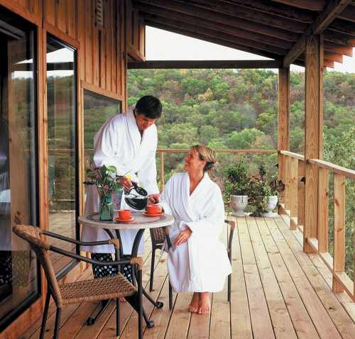 A couple in white robes enjoys coffee on a wooden deck, surrounded by greenery and a peaceful landscape.