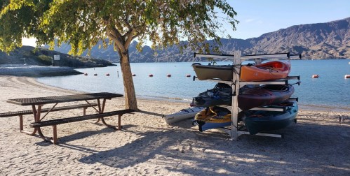 A stack of colorful kayaks by a sandy beach, with a picnic table and mountains in the background.