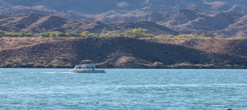 A boat glides across a calm lake, surrounded by rolling hills and mountains in the background.