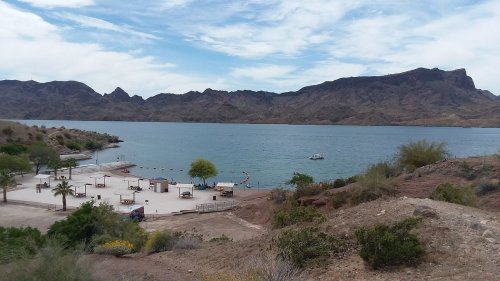 A serene lake surrounded by mountains, with a sandy beach area and a few boats on the water under a cloudy sky.