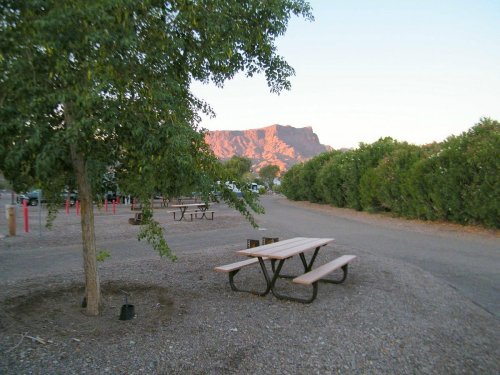 A picnic table under a tree, with a mountain backdrop and a gravel path in a serene outdoor setting.