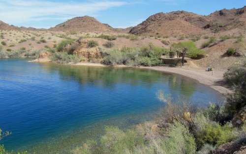 Serene lake surrounded by hills and greenery, with a sandy shore under a clear blue sky.