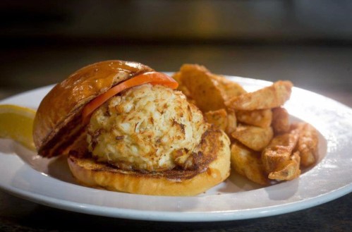 A crab cake sandwich with a tomato slice on a toasted bun, served with seasoned potato wedges and a lemon wedge.