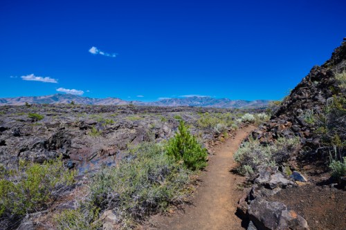 A winding dirt path through rocky terrain and sparse vegetation under a clear blue sky. Mountains are visible in the distance.