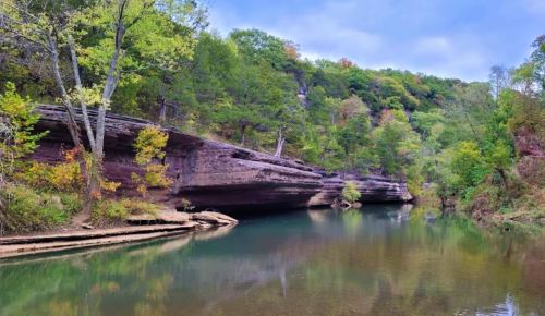 A serene river flows past layered rock formations and lush green trees under a cloudy sky.