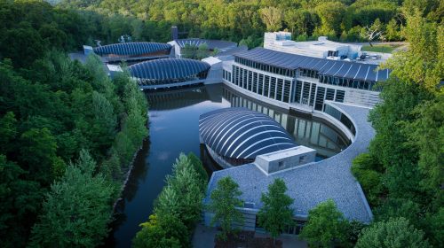 Aerial view of a modern building complex surrounded by trees and water, featuring curved roofs and a natural landscape.