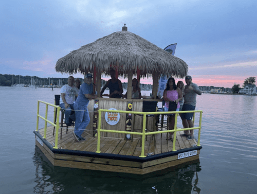 A group of people stands on a floating tiki bar at sunset, surrounded by water and boats in the background.