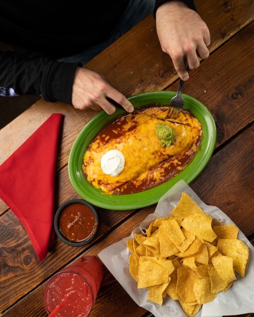 A person is about to cut into a large enchilada topped with cheese, sour cream, and guacamole, with chips and salsa nearby.