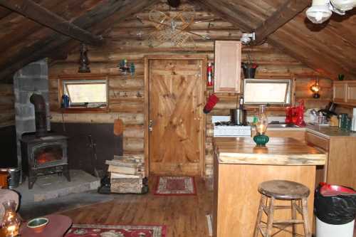 Cozy wooden cabin interior with a kitchen, wood stove, and rustic decor, featuring a door and windows.