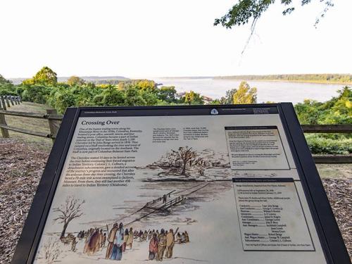 A historical sign titled "Crossing Over" with a river view and illustrations of people by the water's edge.