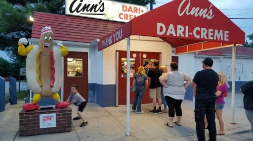 A group of people lines up outside Ann's Dari-Creme, featuring a large inflatable hot dog mascot.