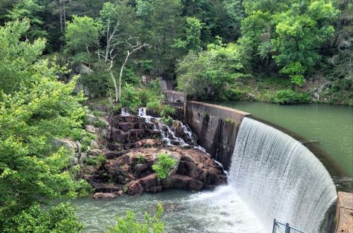 A serene waterfall cascades over a stone dam, surrounded by lush green trees and rocky terrain.