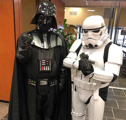 Two costumed characters: one dressed as Darth Vader and the other as a Stormtrooper, posing together indoors.