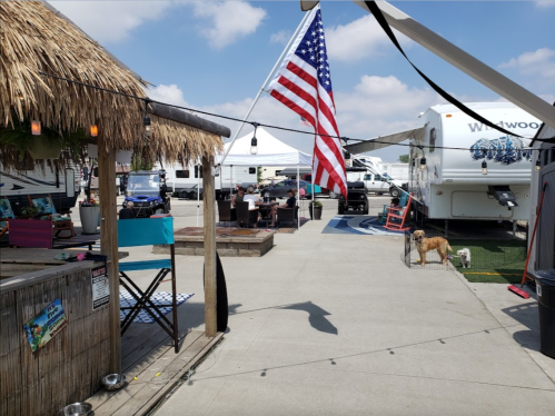 A sunny outdoor area with a tiki hut, RVs, an American flag, and people relaxing, including a dog on the ground.