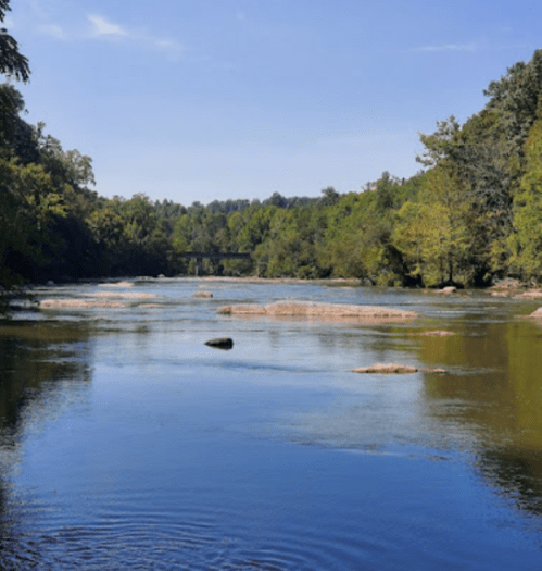 A serene river scene with calm water, rocky outcrops, and lush green trees under a clear blue sky.