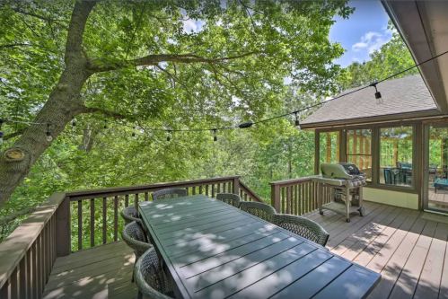A wooden deck with a large table and chairs, surrounded by trees and string lights, overlooking a serene landscape.