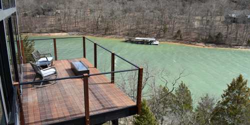 A wooden deck overlooking a green river, surrounded by trees, with a boat docked in the distance.