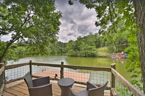 A serene lakeside view from a wooden deck with two chairs and a small table, surrounded by lush greenery.