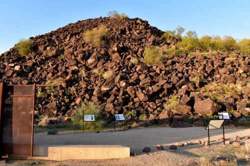 A rocky hill with sparse vegetation, surrounded by informational signs and a gravel path in a desert landscape.