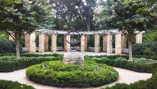 A serene garden with a stone sundial centerpiece, surrounded by lush greenery and benches under a pergola.