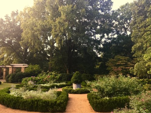 A lush garden with neatly trimmed hedges, colorful flowers, and tall trees under a bright sky.
