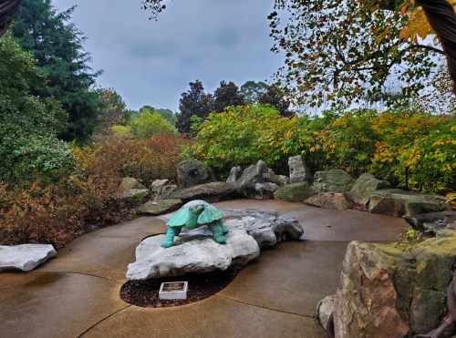 A green turtle sculpture on a rock in a garden surrounded by trees and colorful foliage under a cloudy sky.
