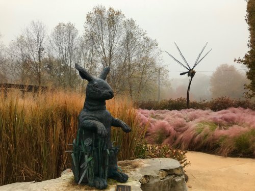 A bronze rabbit statue sits among tall grasses, with a large wind sculpture in the background on a foggy day.