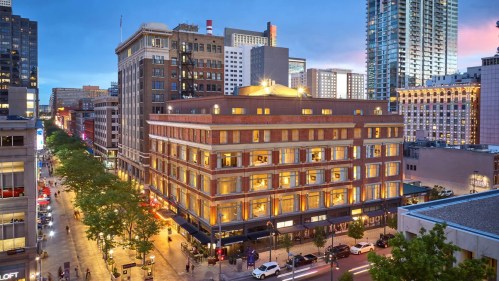 A city street view at dusk featuring a historic building with illuminated windows and modern skyscrapers in the background.