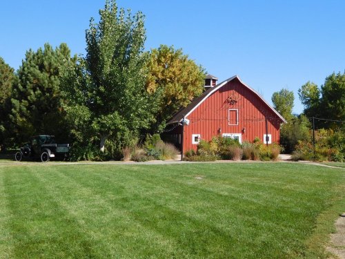 A red barn surrounded by green grass and trees under a clear blue sky. A vintage tractor is parked nearby.