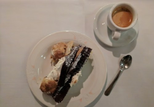 A plate with a slice of cake topped with cream and a scone, alongside a cup of espresso and a spoon.