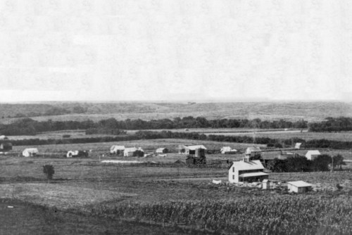 A black and white landscape showing a rural settlement with several buildings and fields under a cloudy sky.