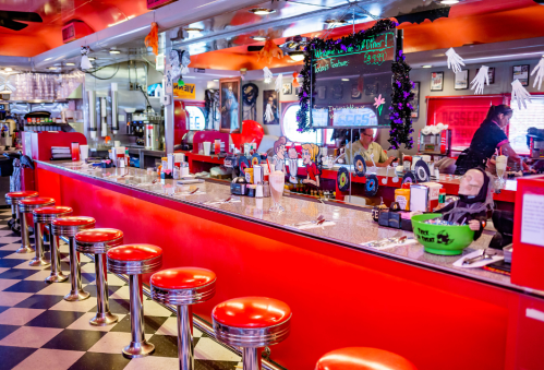 A vibrant diner interior with red stools, a checkered floor, and a festive atmosphere, featuring a counter and decorations.