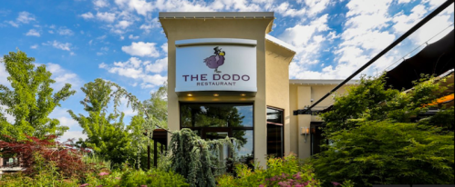 Exterior of The Dodo Restaurant, featuring a sign and lush greenery under a blue sky with scattered clouds.