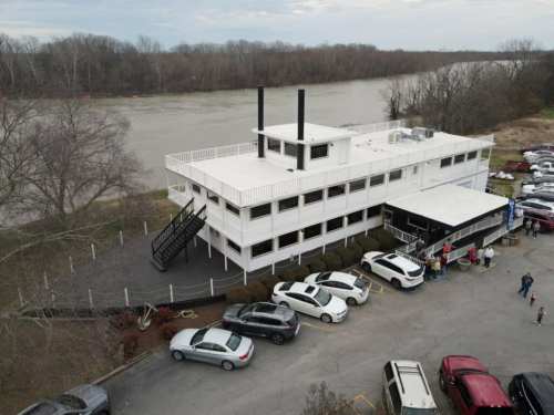 A large white building resembling a riverboat, parked by a river with cars and people nearby.