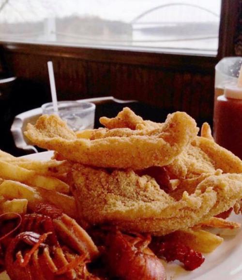 A plate of fried fish, fries, and crawfish, with a scenic view of a bridge in the background.