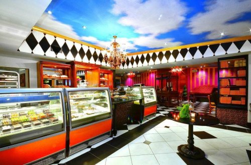 A stylish café interior with a checkered floor, colorful decor, and display cases filled with desserts under a painted sky ceiling.