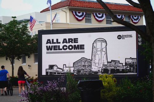 A large sign reads "ALL ARE WELCOME" with a graphic of a water tower, set in a festive outdoor area.