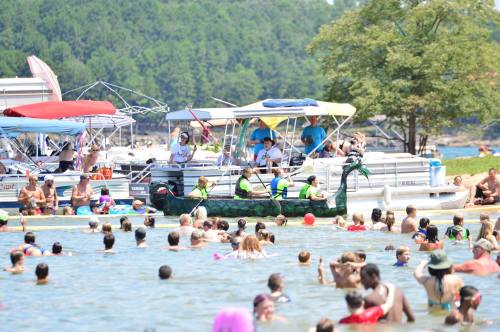 A busy lake scene with people swimming and several boats nearby, enjoying a sunny day.