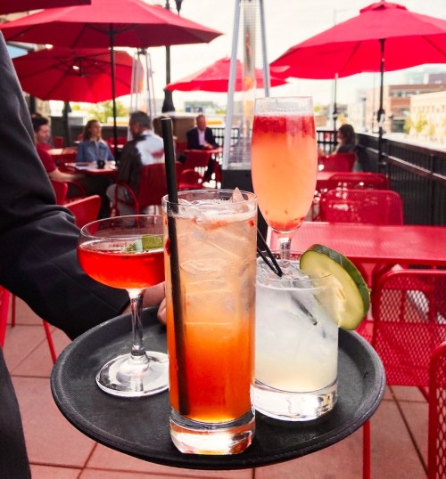 A waiter serves three colorful cocktails on a black tray, with red umbrellas and patrons in the background.