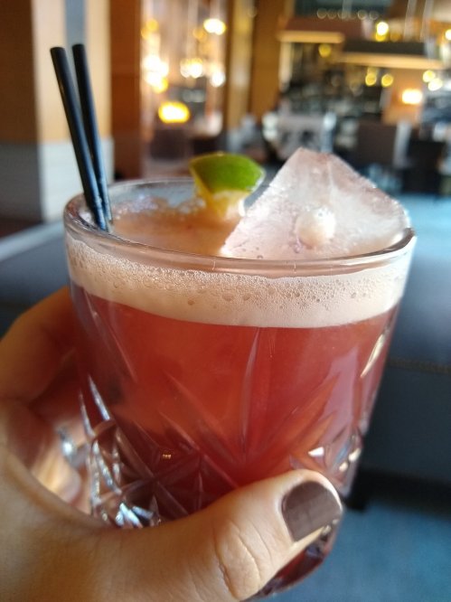 A hand holding a cocktail with ice, a lime wedge, and two straws, set in a stylish bar environment.