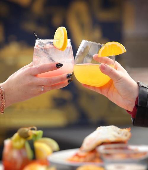 Two hands clink glasses of colorful cocktails, garnished with orange slices, against a blurred background.